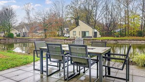 Une terrasse au bord du lac, avec une table et des chaises, entourée d’arbres et avec un petit bâtiment en arrière-plan.