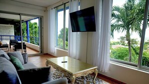 Un salon avec un canapé, une table basse et un téléviseur fixé au mur. Il y a un balcon qui offre une vue sur la verdure environnante.