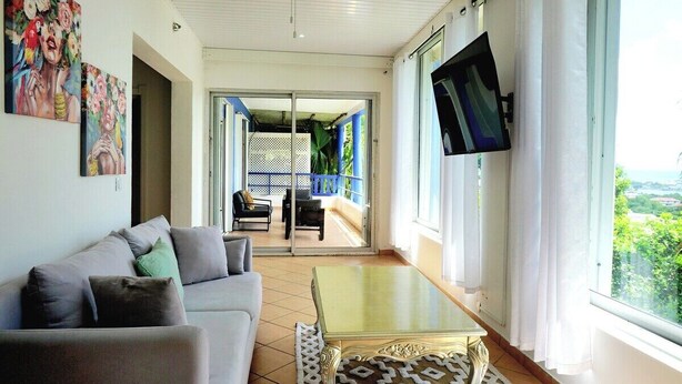Un salon avec un canapé, une table basse et un téléviseur fixé au mur. Il y a un balcon avec vue sur la verdure.