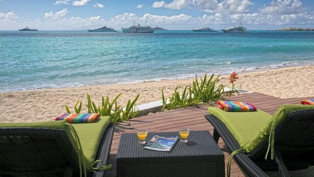 Eine Strandlounge mit zwei Korbstühlen, einem Tisch mit Zeitschrift und Getränken und Blick auf das Meer mit Yachten.