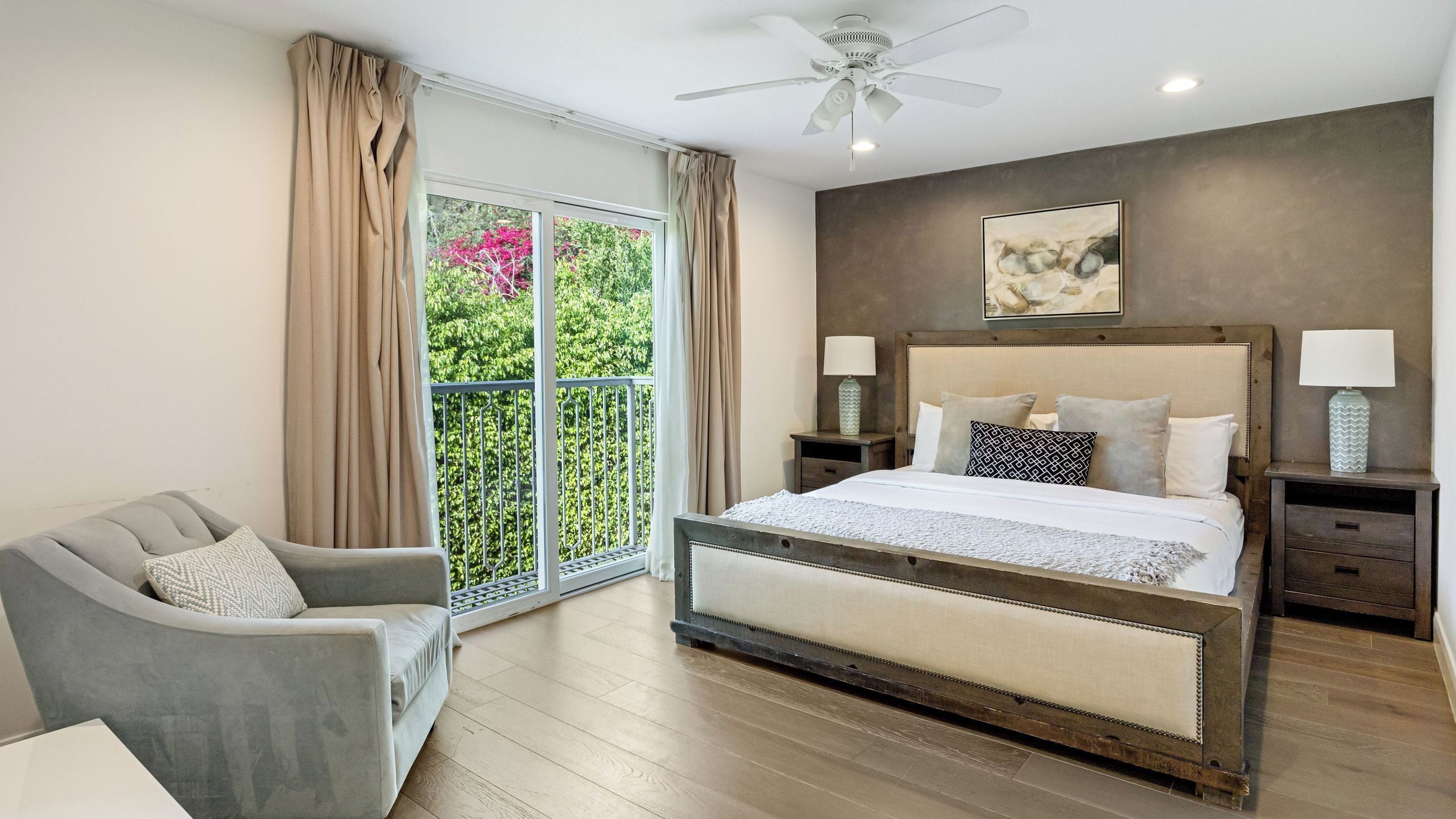 Quarto com cama grande, uma cadeira, mesa de cabeceira, ventilador de teto e janela com cortinas.