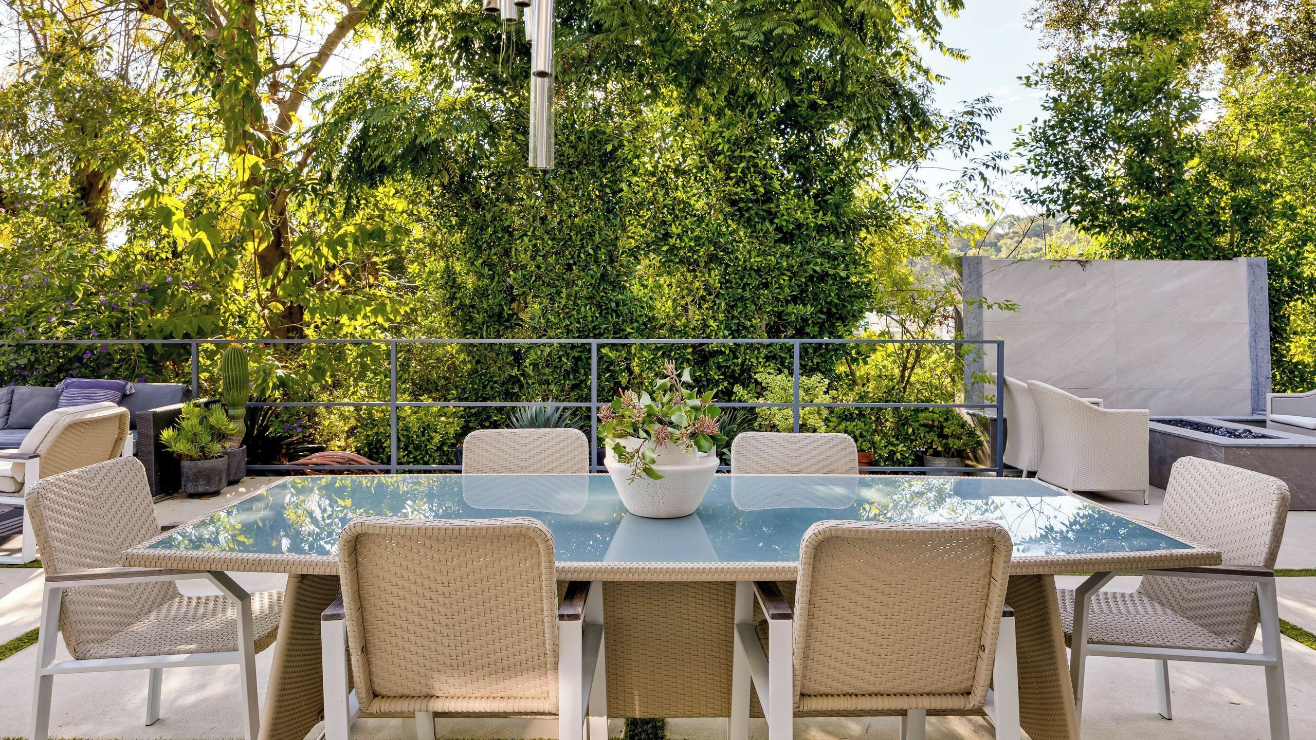 Un patio al aire libre con una mesa de vidrio rodeada de sillas de mimbre, una planta en maceta y una zona de estar con cojines.
