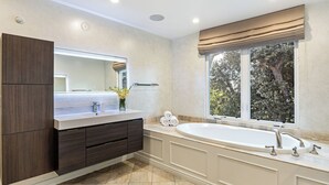 A modern bathroom with a large mirror, dark wood cabinetry, a white vanity, a freestanding bathtub, and a window with a view of trees. - LA Hillside Retreat w/ Pool, Views, Fire Pit & Outdoor Lounging Casa Bellagio by Avantstay (Los Angeles)