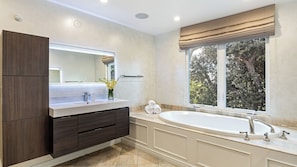 A modern bathroom with a large mirror, dark wood cabinetry, a white vanity, a freestanding bathtub, and a window with a view of trees. - LA Hillside Retreat w/ Pool, Views, Fire Pit & Outdoor Lounging Casa Bellagio by Avantstay (Los Angeles)