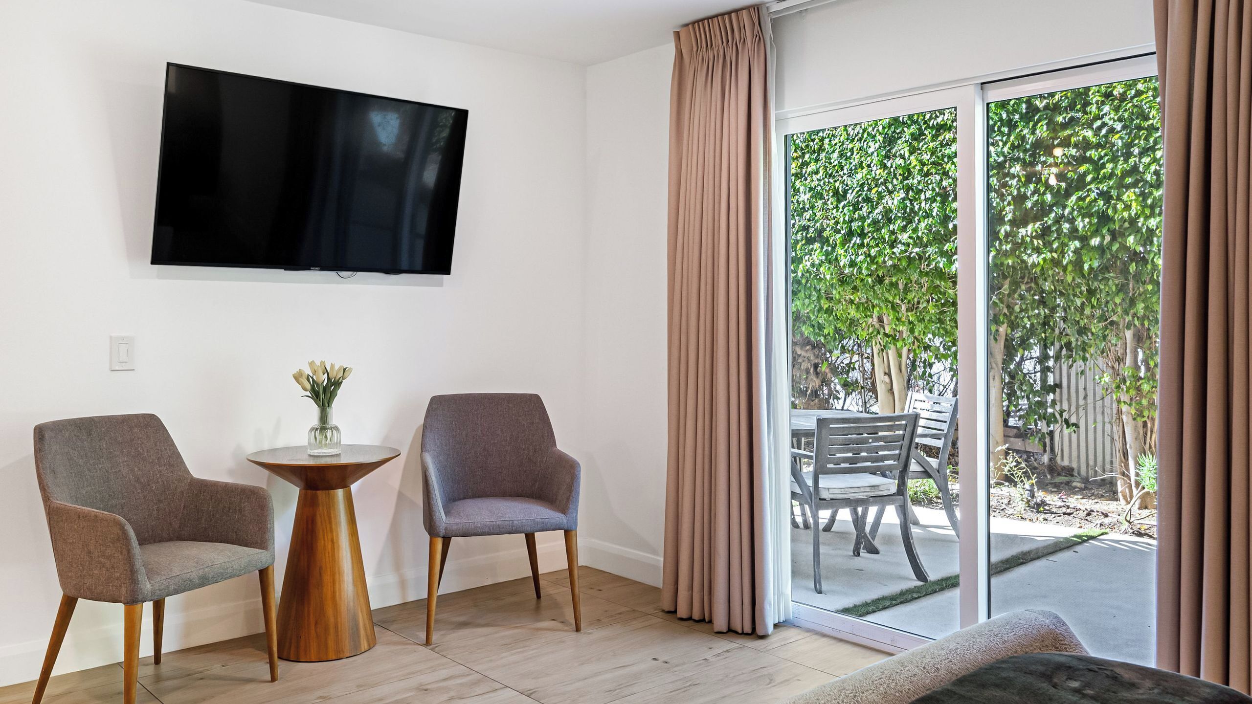 Habitación con televisión de pantalla plana, dos sillones, una mesita de madera con un florero y un ventanal corredero que da a un patio exterior con mesa y sillas.
