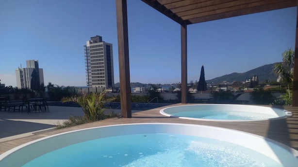 A rooftop pool area with two hot tubs, wooden deck, and a view of city buildings and mountains. - Skyline Confort Club - Blue Horizon (Penha)