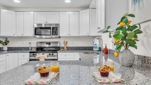 Een moderne keuken met witte kasten, een RVS magnetron, een dubbele oven en een granieten aanrecht. Op het aanrecht staan twee muffins, een glas sinaasappelsap en een vaas met citroenen.