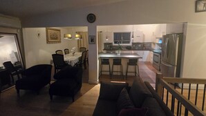 A modern living room with a dark-colored sofa and a dining area with a table and chairs. A kitchen with a refrigerator and a window is visible in the background. - Nature view retreat (Gatineau)