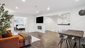 A modern living room with a sofa, a dining table with chairs, a flat-screen TV mounted on the wall, and a large mirror. - Bright, New & Spacious 1 Bedroom Suite! Family friendly & central to shopping. (Burnaby)