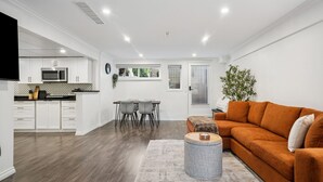 A modern kitchen with white cabinets, a dining area with a table and chairs, and a living area with a sofa and a rug. - Bright, New & Spacious 1 Bedroom Suite! Family friendly & central to shopping. (Burnaby)