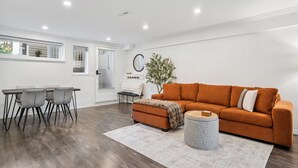 A modern living room with a wooden dining table, grey chairs, a brown sofa, and a grey rug. - Bright, New & Spacious 1 Bedroom Suite! Family friendly & central to shopping. (Burnaby)