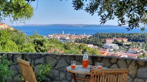 Eine Steinswand mit zwei Holzstühlen und einem Tisch, mit Blick auf eine Küstenstadt mit Gebäuden und blauem Meer.