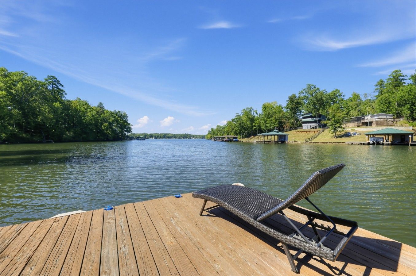 A wooden dock with a lounge chair facing a calm river, surrounded by greenery and buildings.