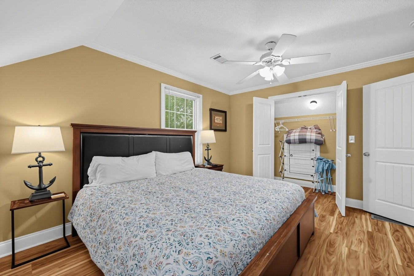 A bedroom with a bed, two bedside tables with lamps, a ceiling fan, a window with blinds, and a door leading to a closet with hanging clothes.