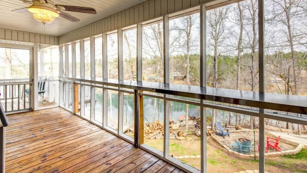 A covered porch with wooden flooring and large windows offering a view of a wooded area and a body of water.