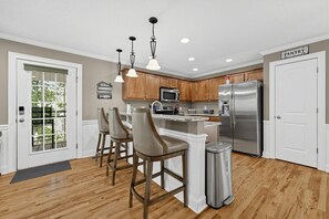 A kitchen with wooden cabinets, a stainless steel refrigerator, a microwave, and a bar area with stools. - Lake Sinclair 4BR | Private Dock & Game Room (Sparta)