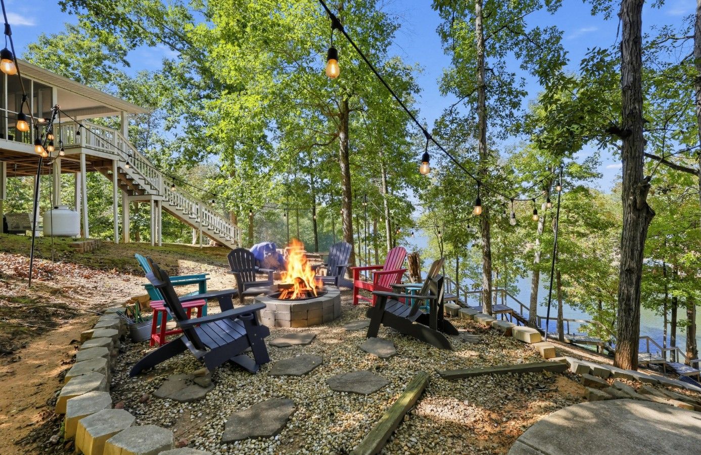 An outdoor seating area with Adirondack chairs around a fire pit, a wooden deck, and a view of a lake.