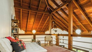 A high-ceilinged room with wooden beams, a bed with colorful pillows, a small table with a plant, and a chair. - Kite House - Ibiraquera - SC (Imbituba)