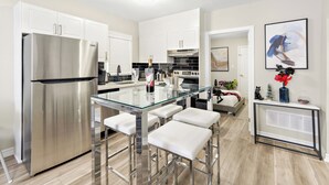Une cuisine moderne dotée d’un réfrigérateur en acier inoxydable, d’un îlot central avec une plaque de cuisson en verre et des tabourets de bar, de meubles blancs et d’un dosseret en carreaux noirs.