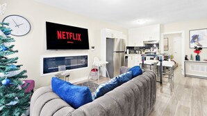 Un salon moderne avec un canapé gris, un téléviseur à écran plat, un sapin de Noël et une cuisine en arrière-plan.