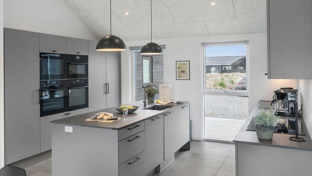 Une cuisine moderne dotée d’un îlot central, de fours encastrés, de luminaires suspendus et offrant une vue sur l’extérieur.