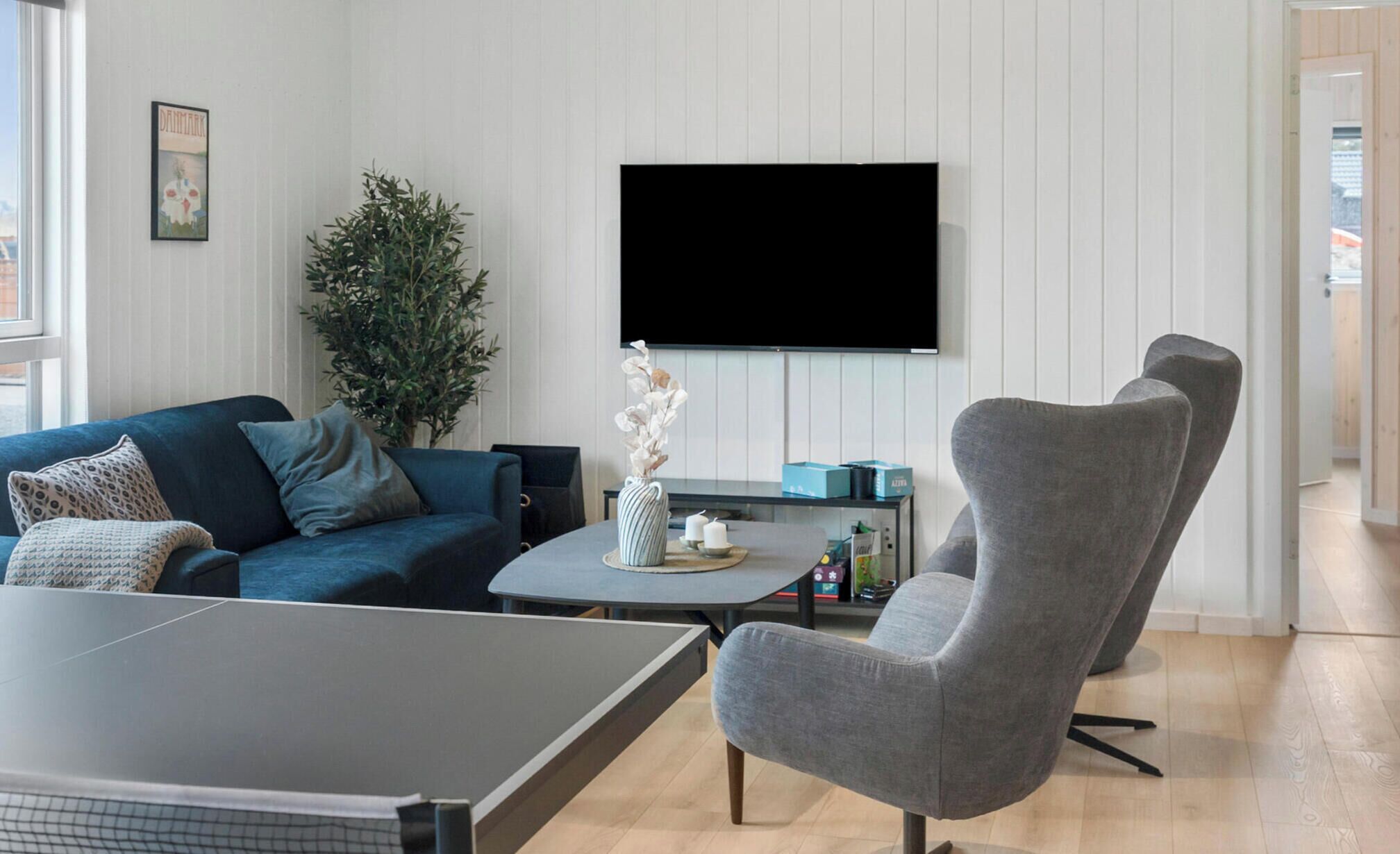 Ein Wohnzimmer mit einer Couch, einem Couchtisch, einem grauen Sessel, einem flachbildfernseher an der Wand und einer Topfpflanze.