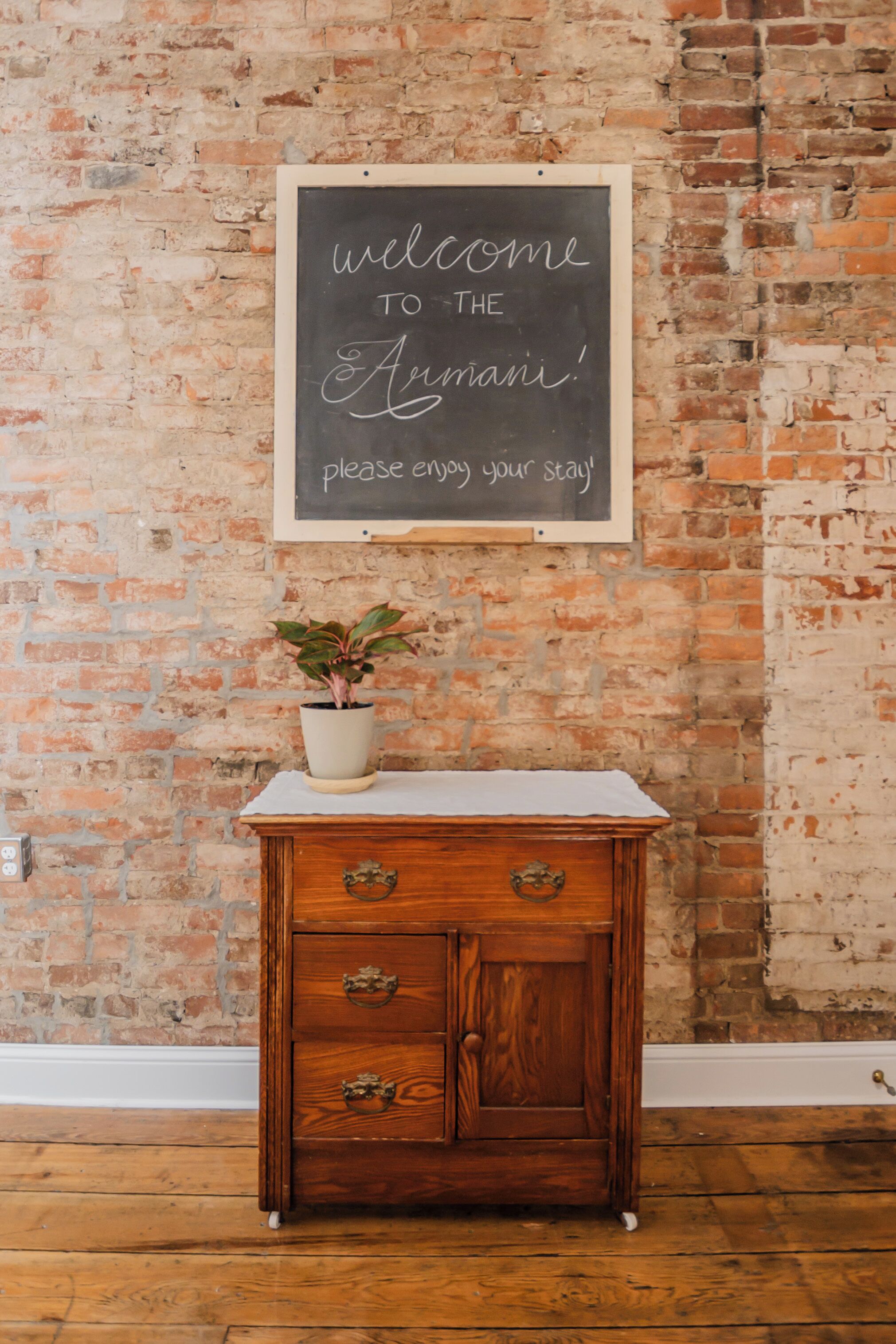 A wooden cabinet with drawers and a potted plant on top, placed against a brick wall with a chalkboard sign above it.