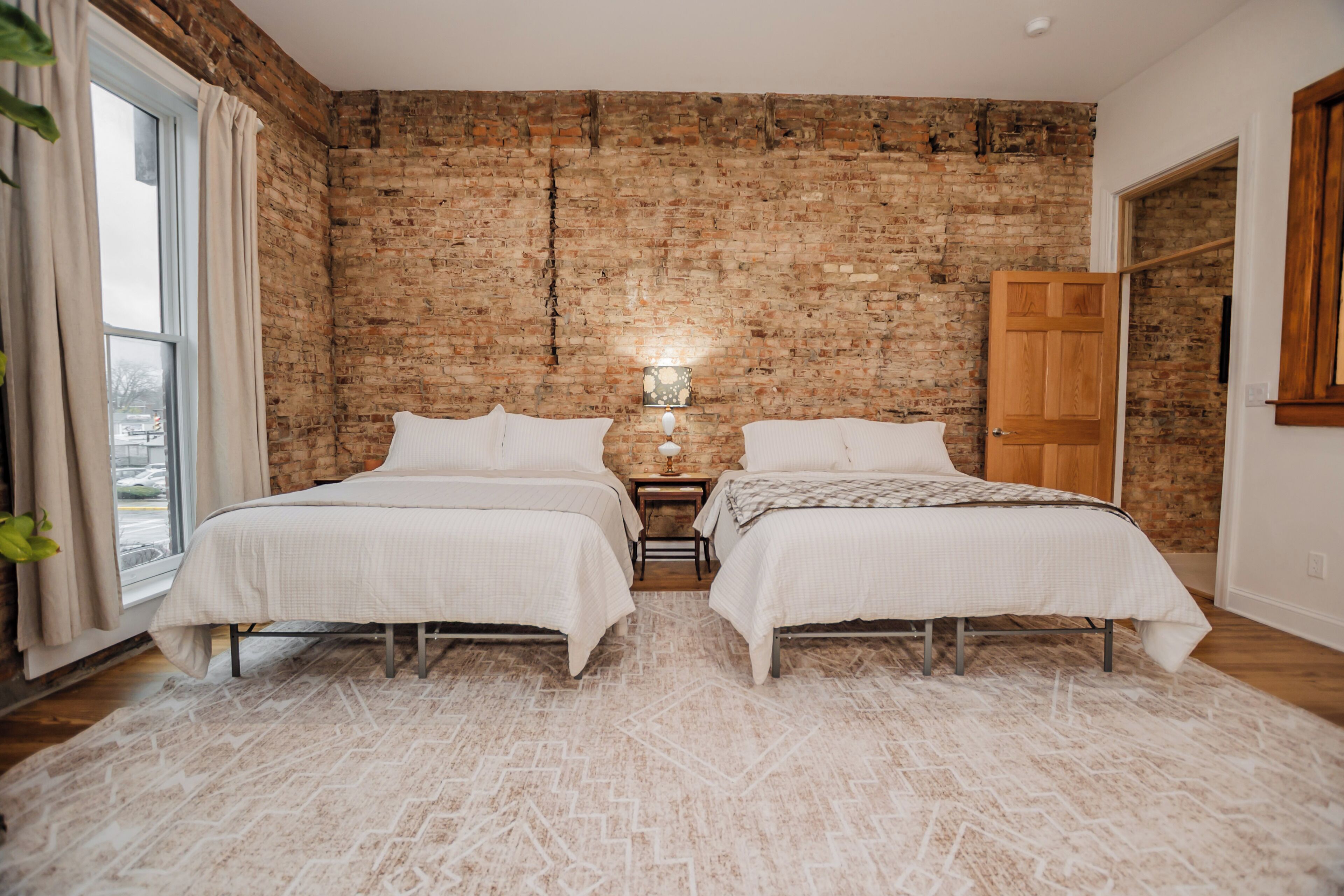 A bedroom with two beds, a brick wall, a window with curtains, and a wooden door.