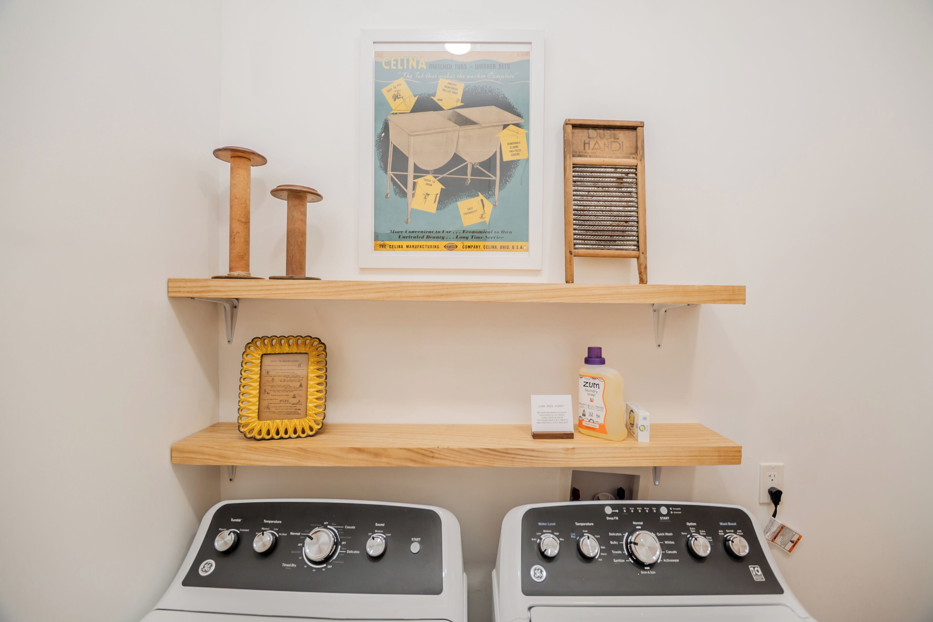 Une buanderie équipée de deux machines à laver, de rayonnages en bois où sont rangés divers objets, et une affiche accrochée au mur.
