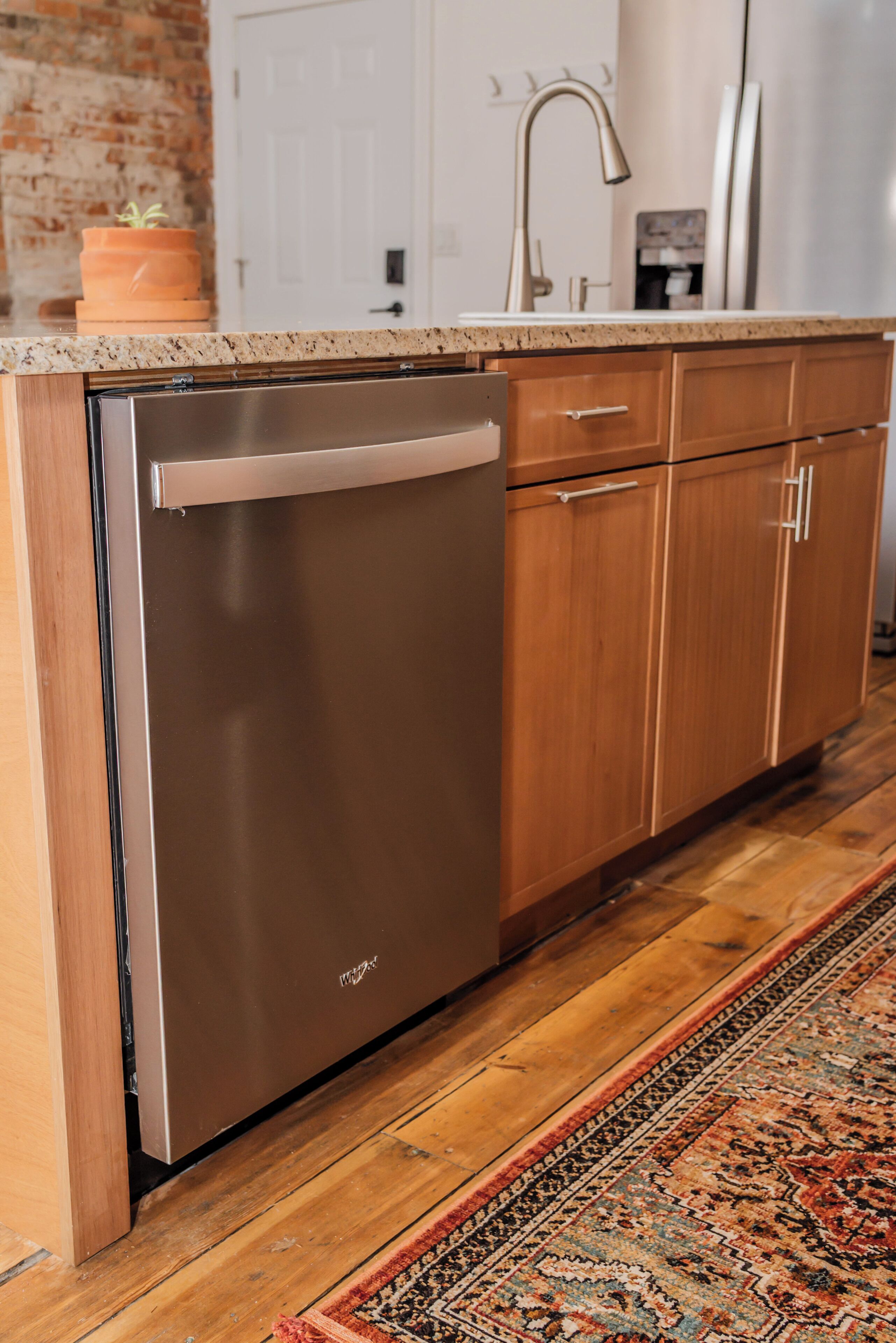 A kitchen with a stainless steel dishwasher, wooden cabinets, a granite countertop, and a patterned rug.