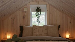 Un dormitorio acogedor en el ático, con techo inclinado, una cama con dos almohadas, una planta colgante y dos lámparas de noche.