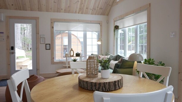 Una habitación con una mesa redonda de madera, sillas blancas, un centro de mesa tejido y ventanas grandes.