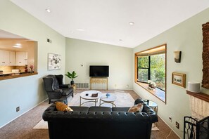 A living room with a black leather sofa, a coffee table, a television, and a large window with a view of greenery. - Mid-Century Escape. Hot Tub. Lush Garden. A/C. Bar (Ojai)