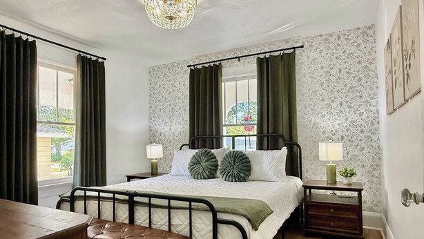 Ein Schlafzimmer mit einem Bett, zwei Nachttischen, einer Bank, einer Chandelierleuchtre und einem Fenster mit Vorhängen.