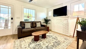 Ein Wohnzimmer mit braunem Sofa, einem Holztisch, einem gemusterten Teppich, einem Deckenventilator, einem an der Wand montierten Fernseher und einem Kamin aus weißen Ziegelsteinen.