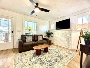 A living room with a brown sofa, a wooden coffee table, a patterned rug, a ceiling fan, a TV mounted on the wall, and a white brick fireplace. - 3 King Beds -Fast WiFi, Fenced Yard & Pet Friendly (Lakeland)