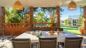 Un espacio de comedor al aire libre techado con una mesa dispuesta para dos personas, rodeado de pilares de ladrillo y vistas a una piscina y follaje.