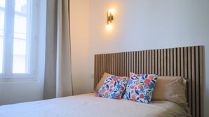 Una cama bien hecha con almohadones florales, cabecera de listones de madera y lámpara fijada a la pared.