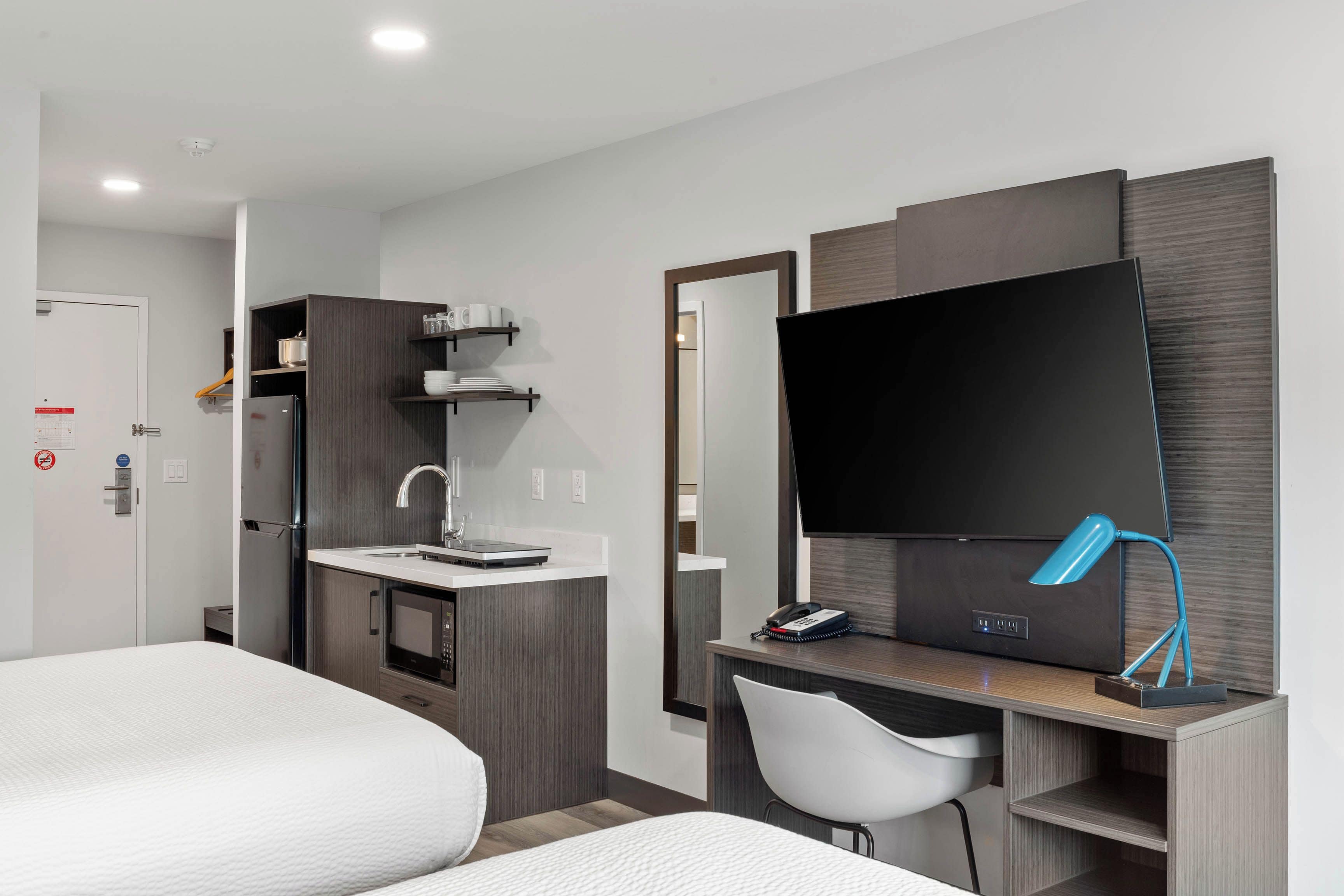 Ein Hotelzimmer mit Bett, Schreibtisch mit Stuhl, Flachbildfernseher, Spiegel und einer Küchenzeile inklusive Kühlschrank und Mikrowelle.
