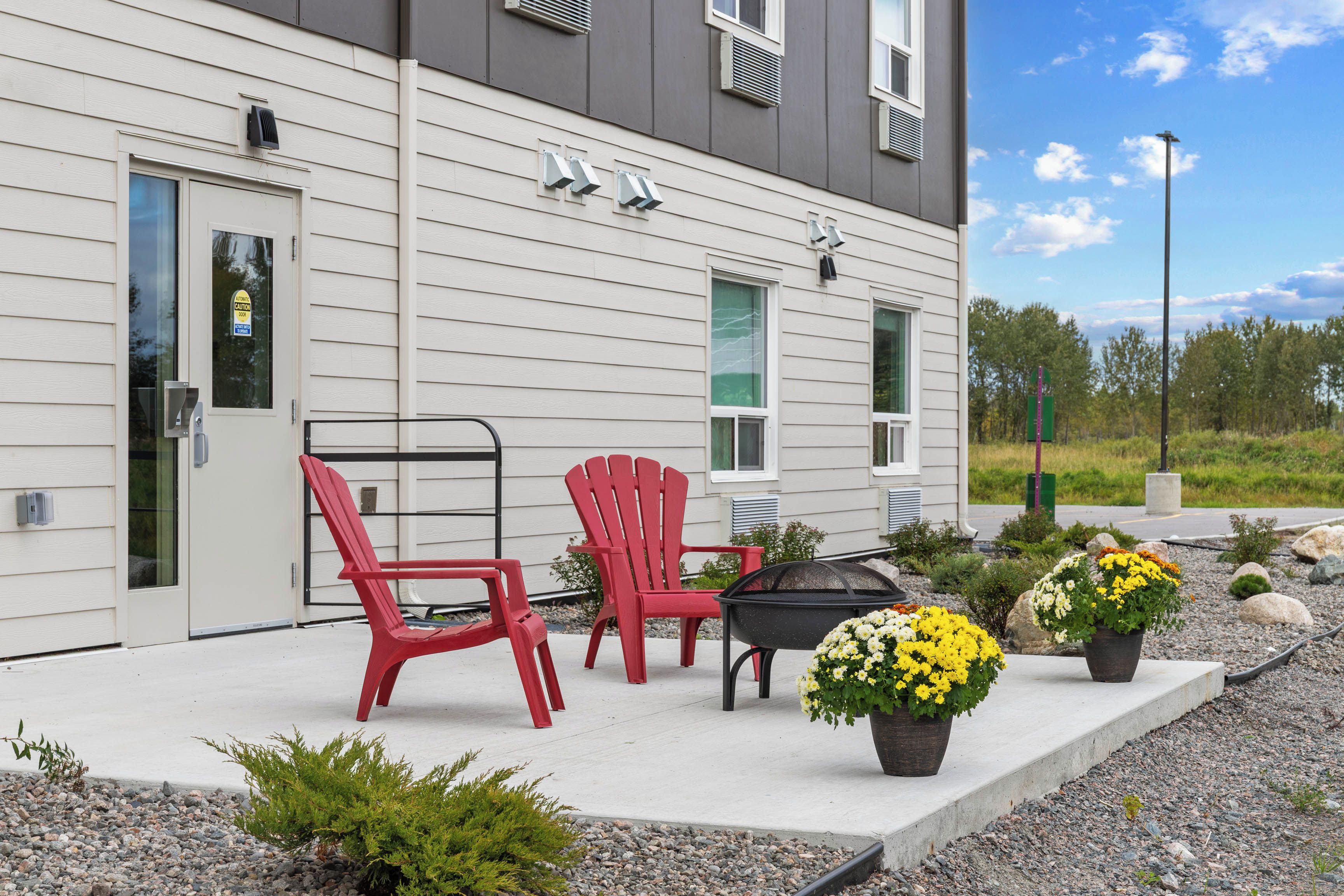 A building with a grey exterior, a door, and windows. There are two red chairs and a black table on the porch.