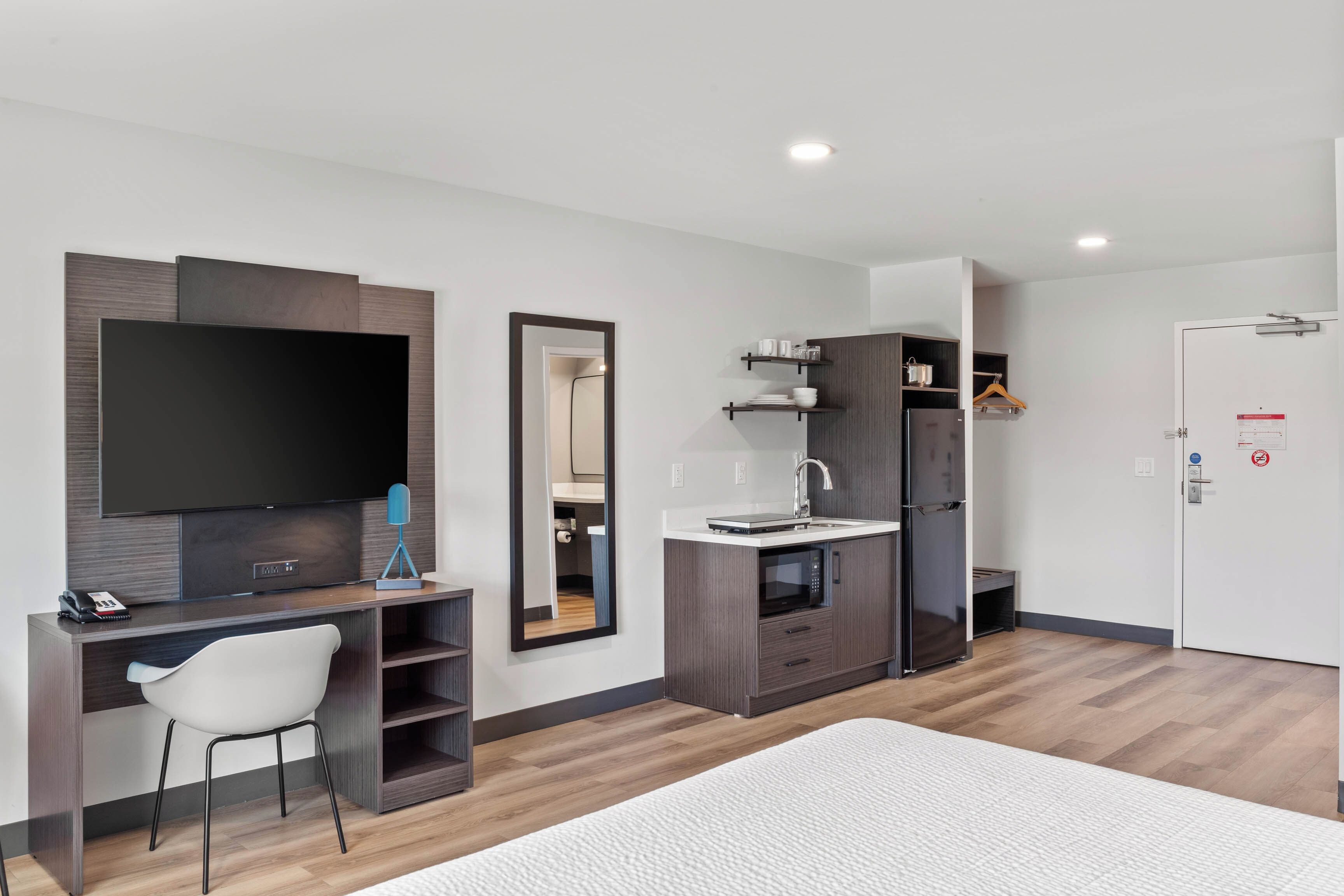 Una habitación de hotel moderna con una televisión de pantalla plana, un escritorio con silla, una cocina integrada con microondas y un armario.