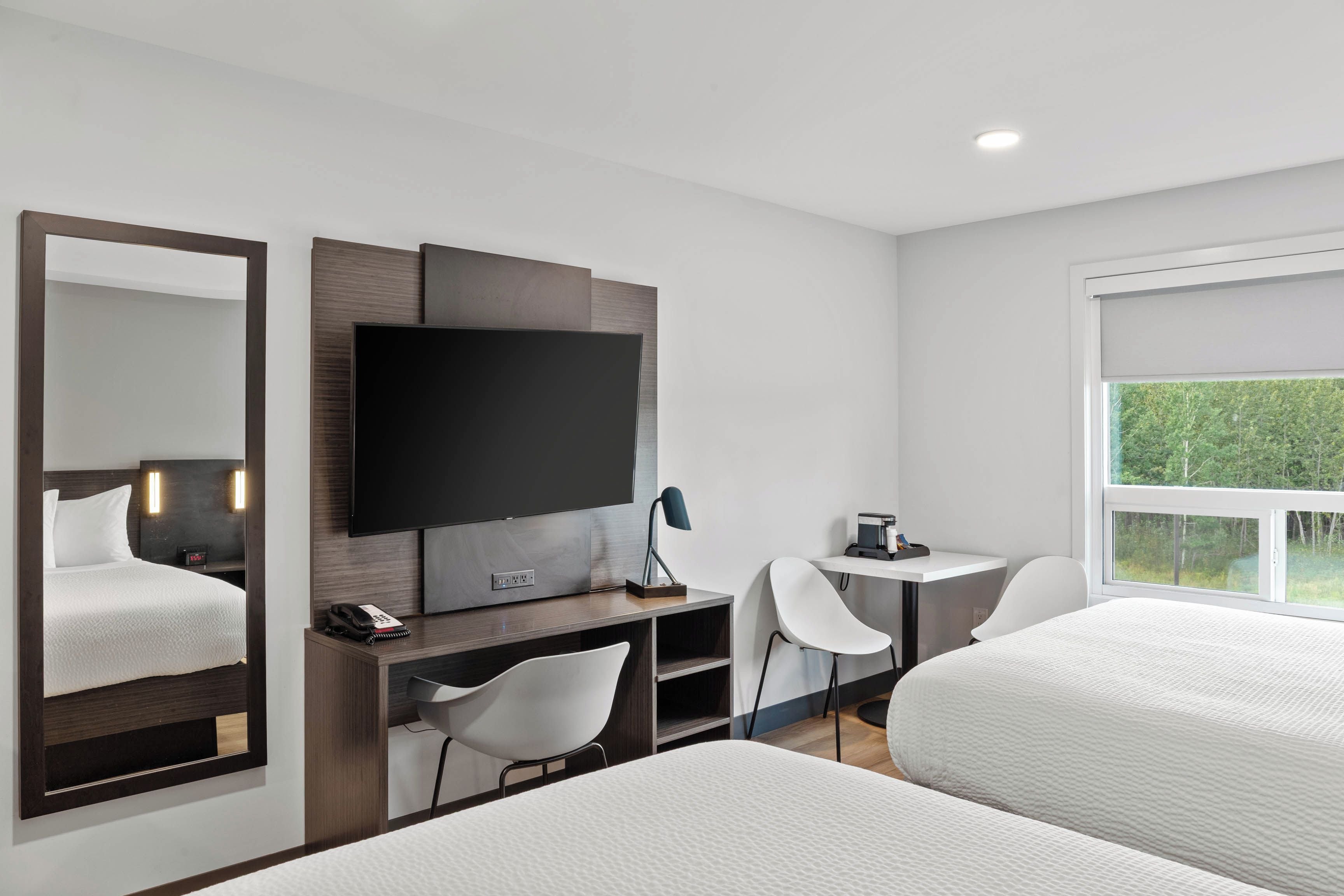A modern hotel room with a large flat-screen TV, a desk with a chair, a small table with a coffee maker, and two beds.