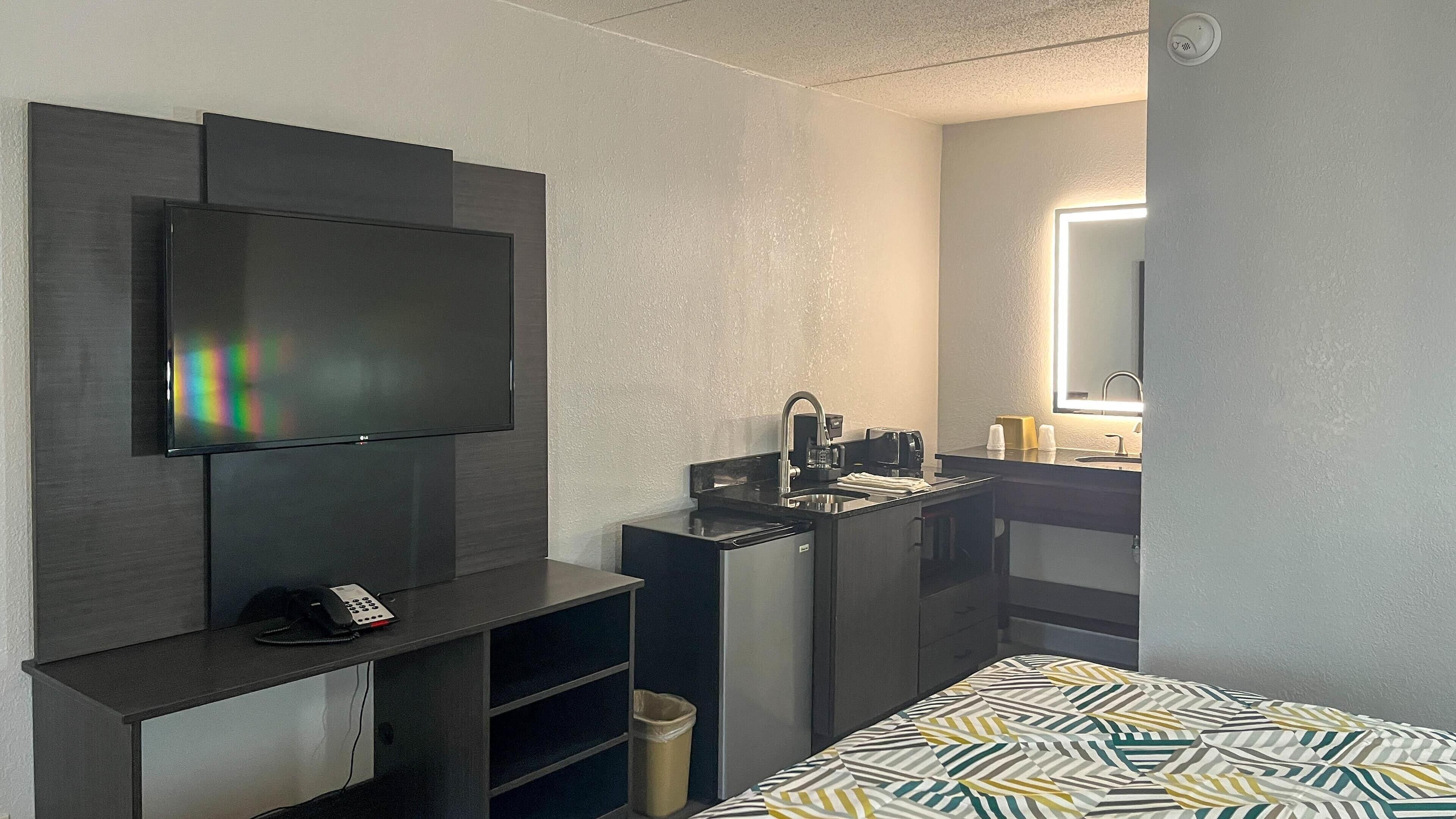 A compact hotel room with a flat-screen TV mounted on the wall, a small kitchenette with a sink, and a bed with a patterned bedspread.