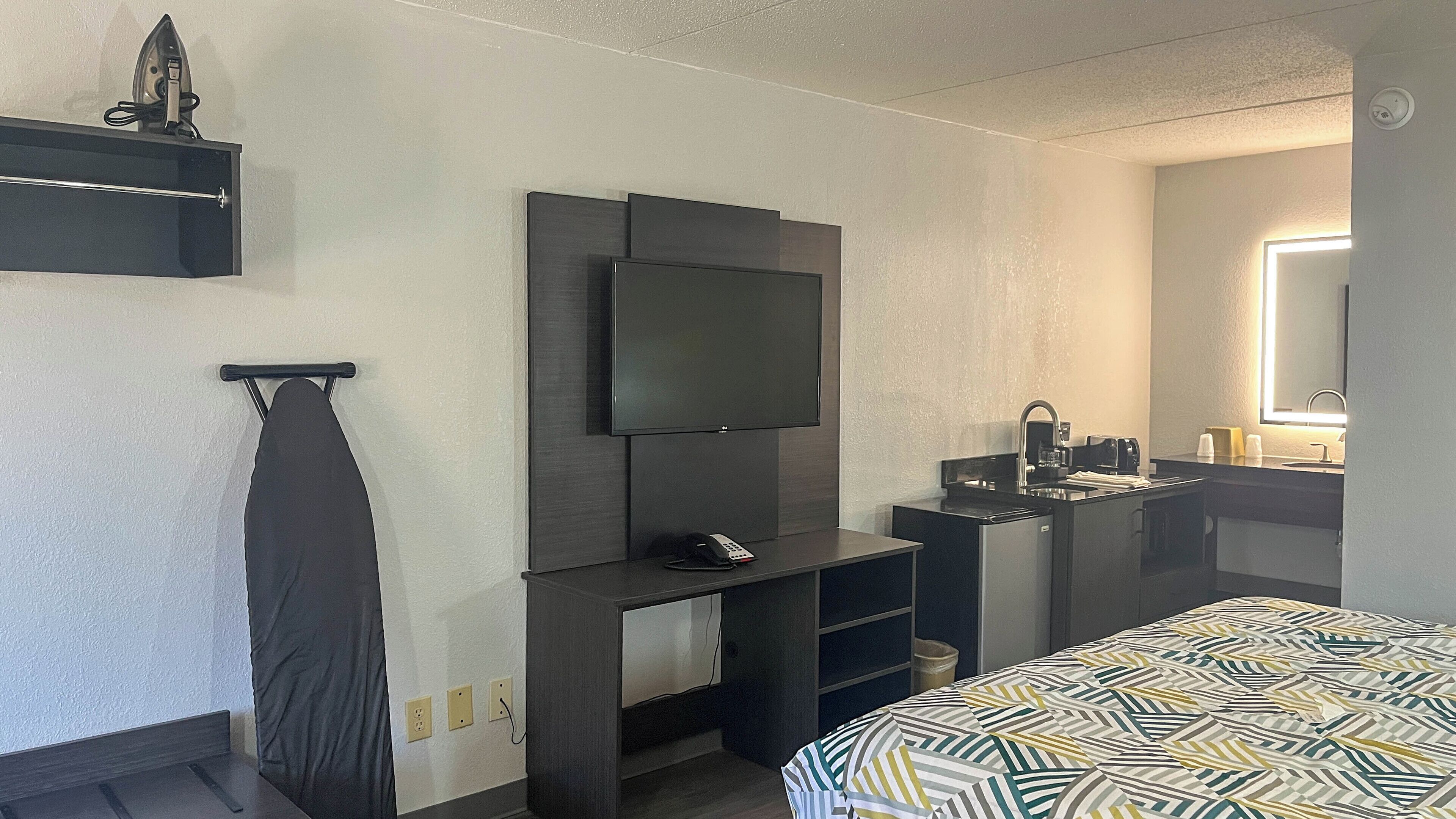 Habitación de hotel con televisor de pantalla plana en la pared, un escritorio pequeño con control remoto, una cama con colcha estampada, un lavamanos con espejo e implemento para planchar colgado del techo.