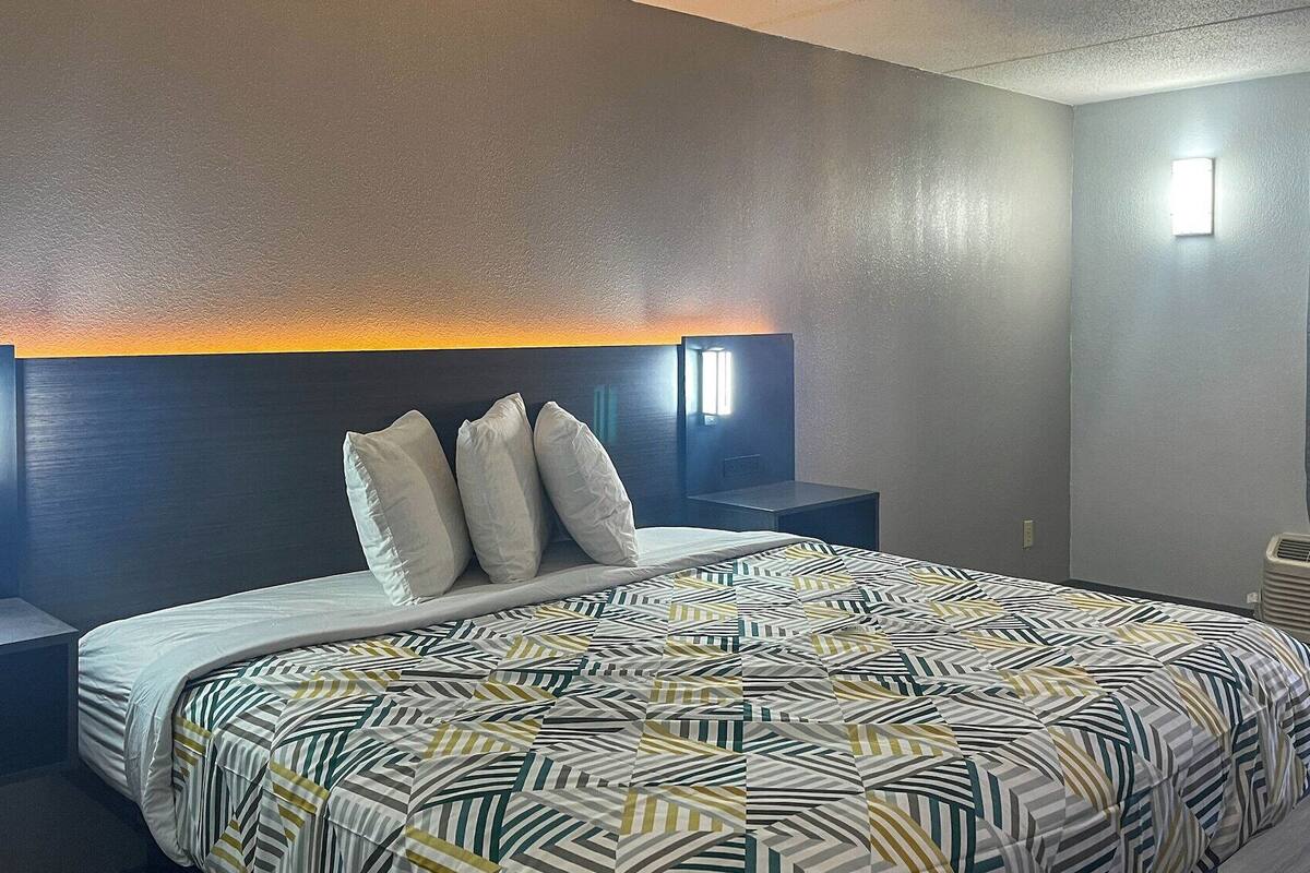 A hotel room with a large bed, two bedside tables, a headboard with built-in lighting, a window with curtains, and a wall-mounted air conditioner.