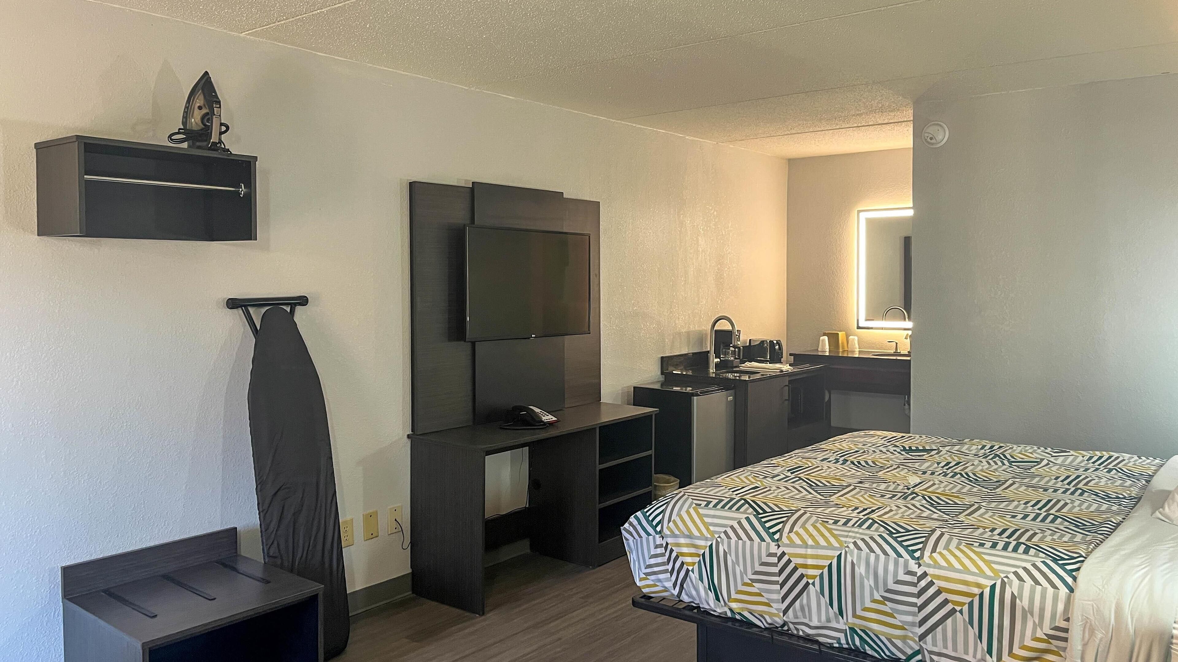 Una habitación de hotel con cama, televisión, escritorio con silla, kitchenette pequeña y baño con espejo.