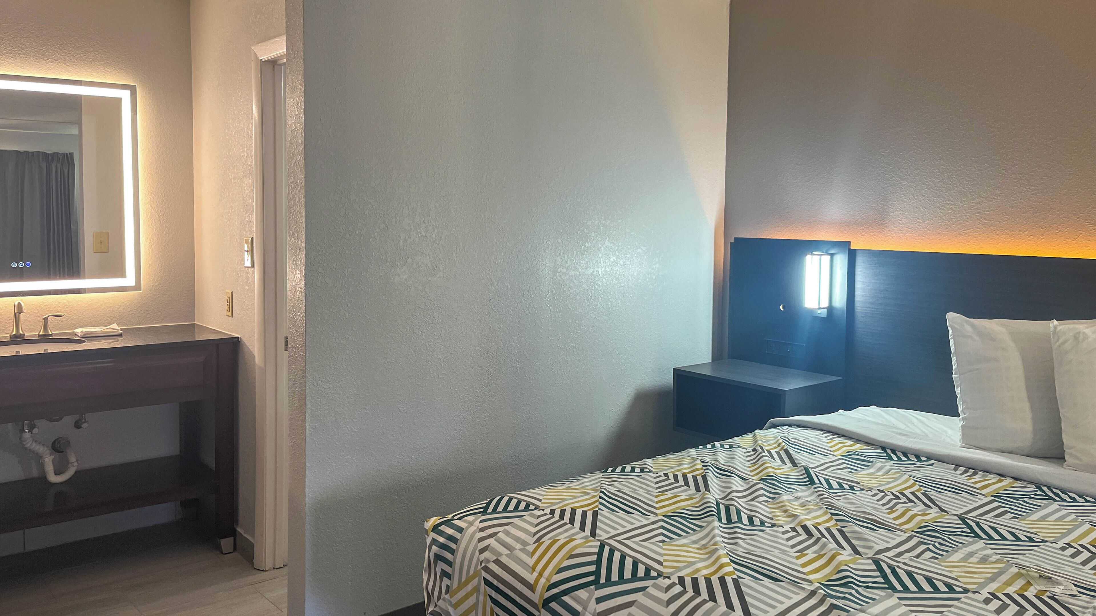 A hotel room with a bed, a bedside table, a nightstand, a mirror, and a wall-mounted light fixture.