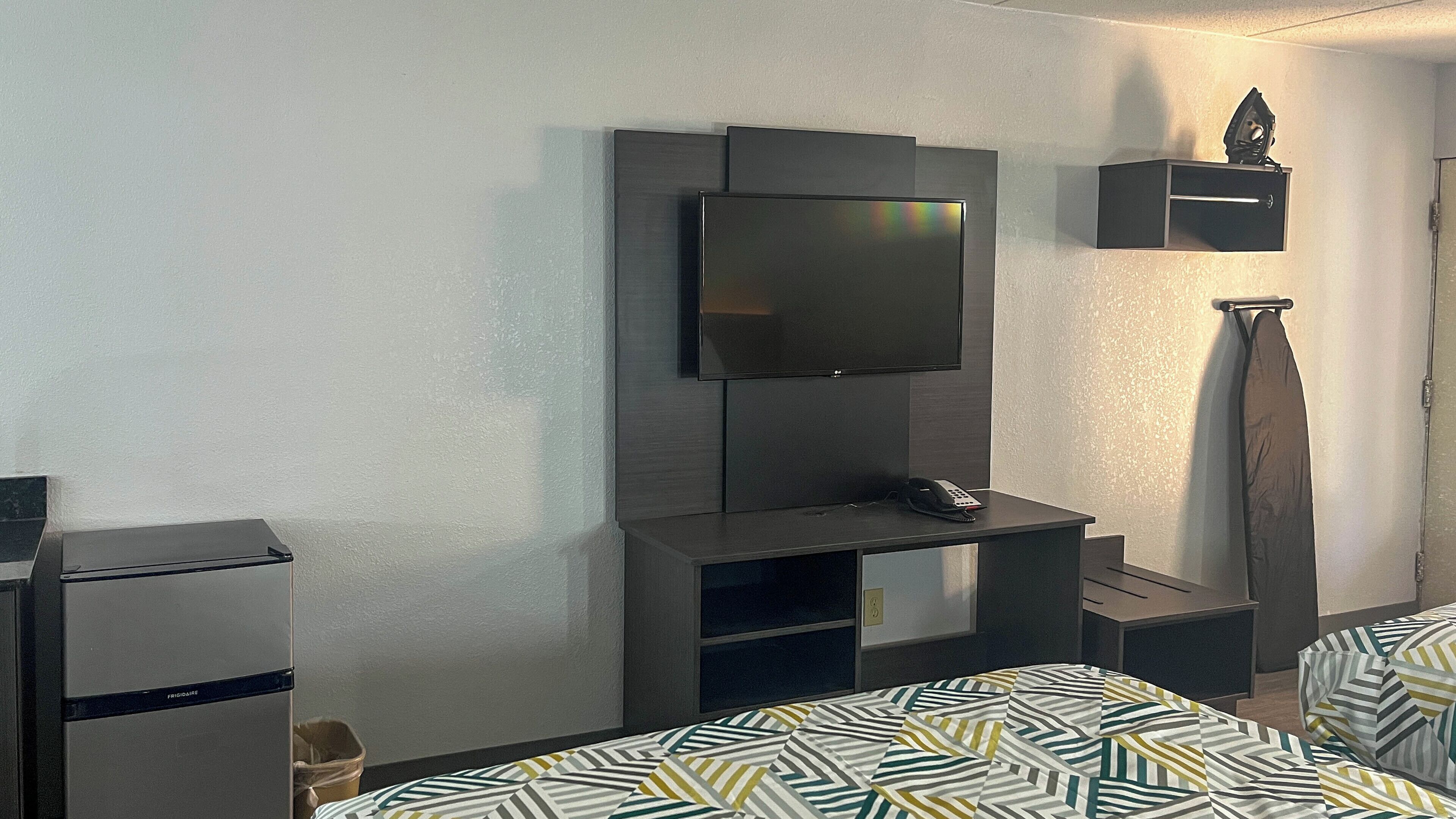 Habitación de hotel con televisor de pantalla plana en la pared, un escritorio pequeño con un teléfono, un refrigerador minibar y una cama con ropa de cama estampada.