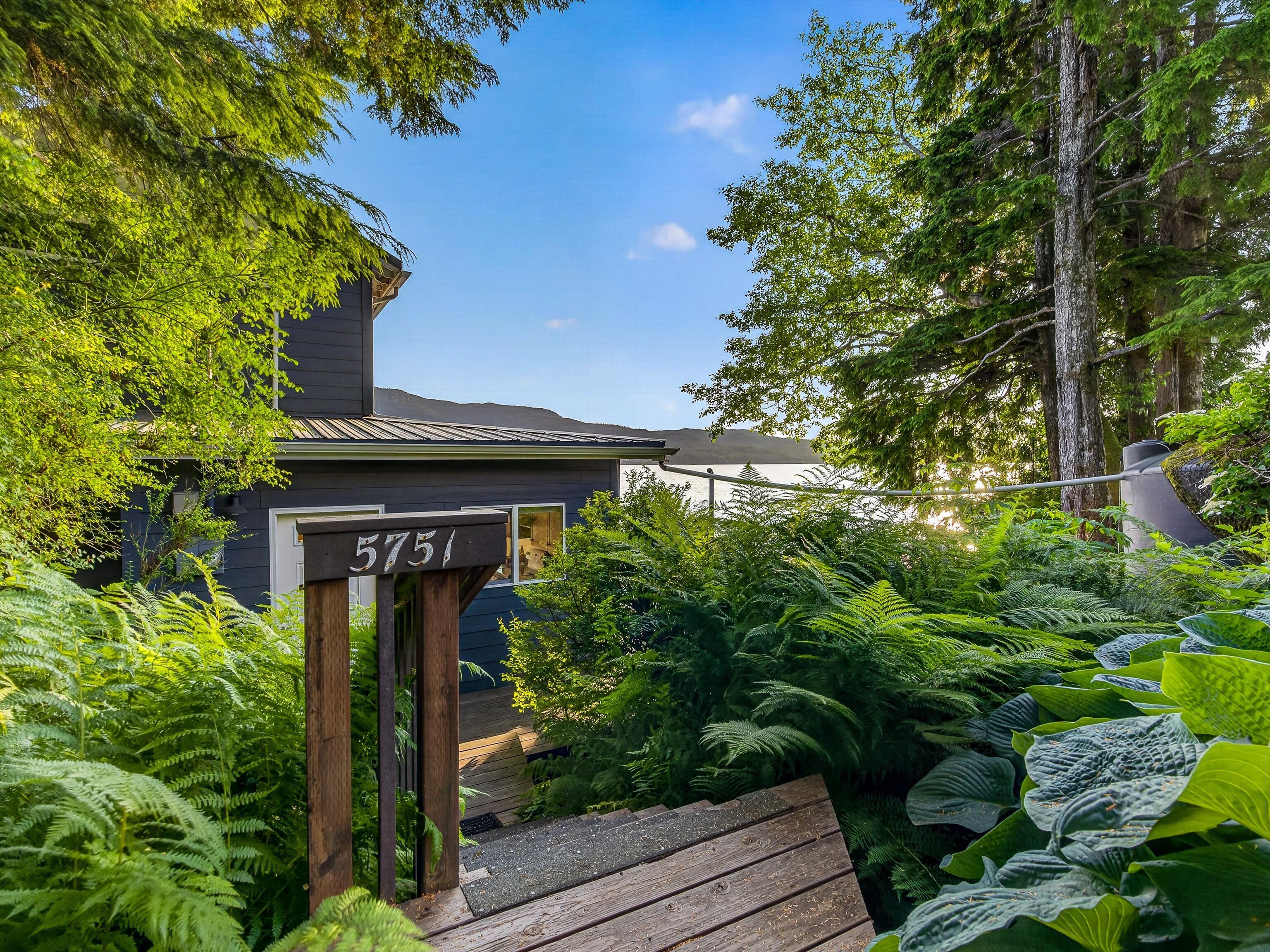 A house with a wooden number sign displaying '5751' in front of a lush garden with various plants and a clear sky in the background.