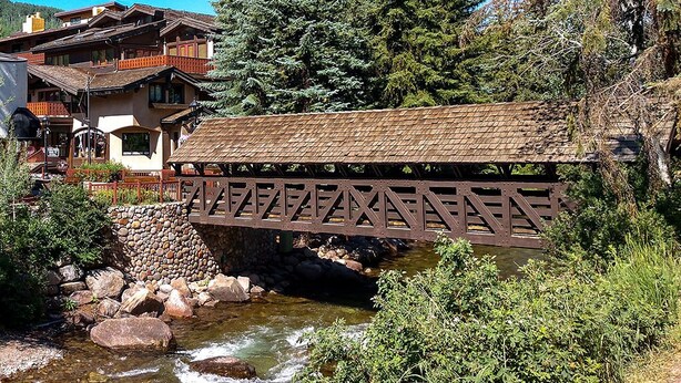 A wooden covered bridge over a stream with a building in the background. - Cozy 1-bedroom condo seconds from bus line in East Vail, perfect for getaways. (Vail)